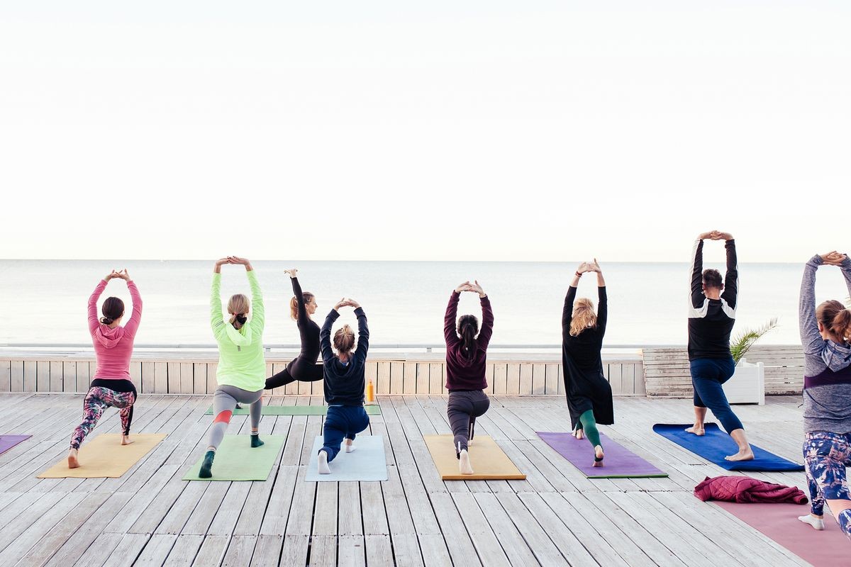 Group of sporty people doing stretching exercise as trainer helps in yoga lesson at wooden fitness terrace on the beach. Teacher assists to make warrior pose, Yoga Practice Exercise Class Concept